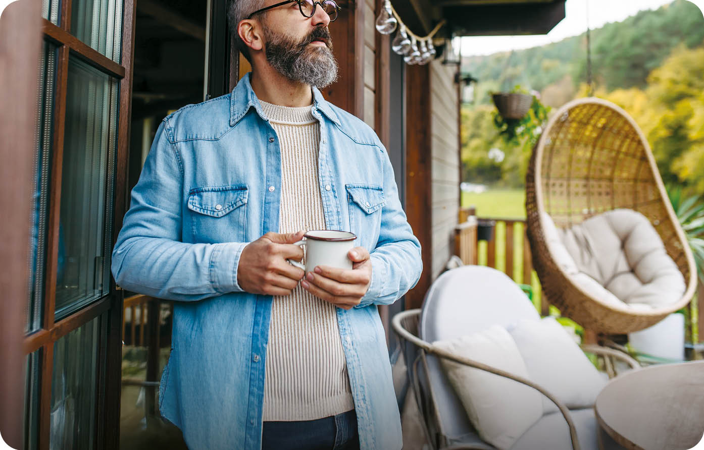 Man is enjoying cup of warm tea, coffee, having relaxing moment at home, standing on patio and looking at autumn nature around
