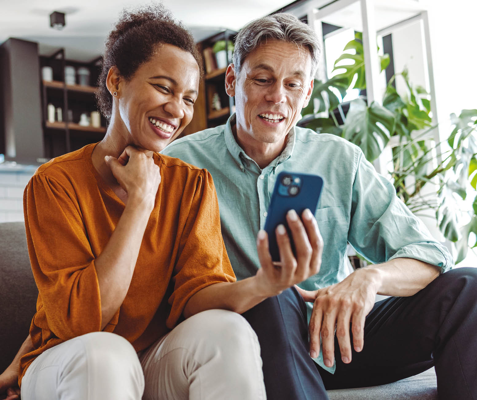 A beautiful African-American woman and a good-looking man sitting in the living room. They are looking at the smart phone and smiling