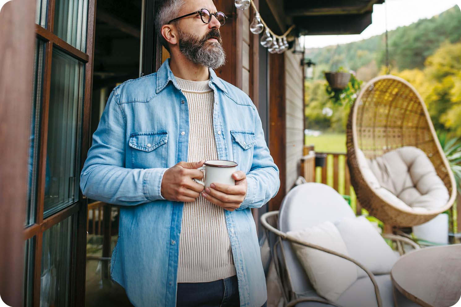 Man is enjoying cup of warm tea, coffee, having relaxing moment at home, standing on patio and looking at autumn nature around