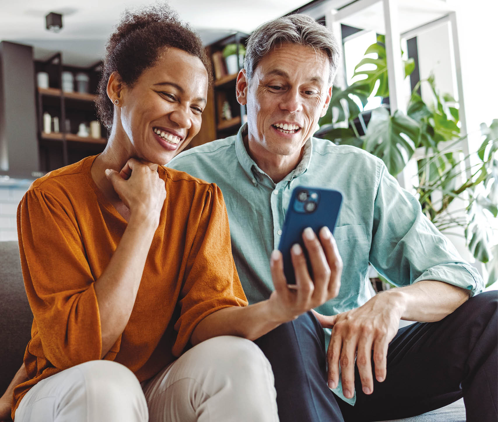 A beautiful African-American woman and a good-looking man sitting in the living room. They are looking at the smart phone and smiling