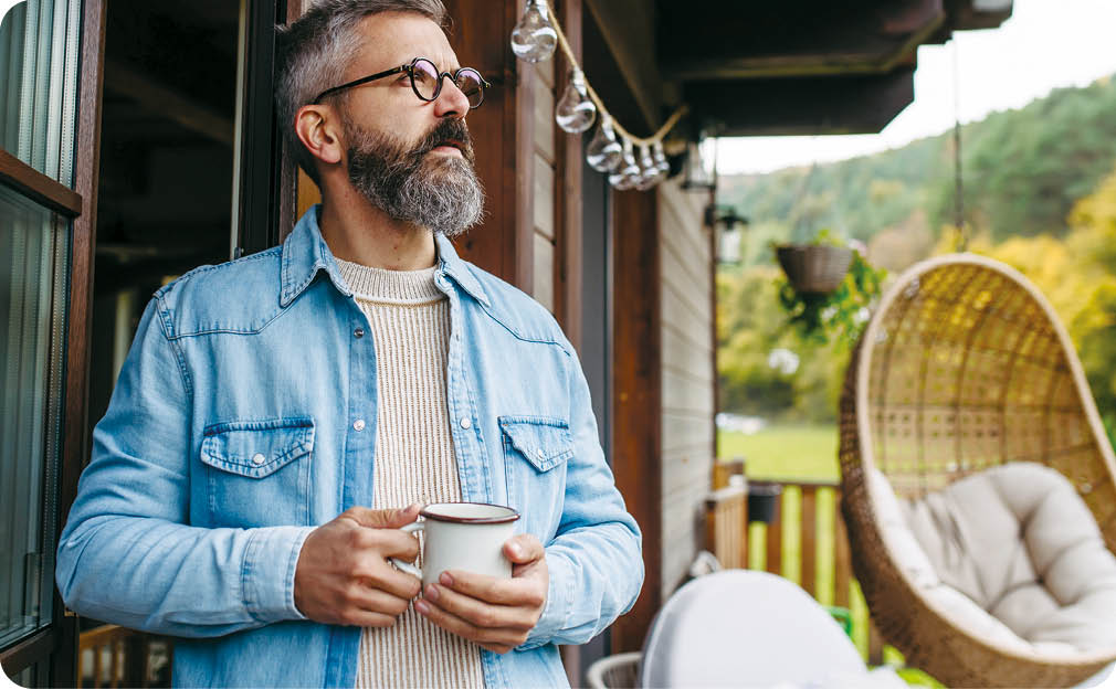 Man is enjoying cup of warm tea, coffee, having relaxing moment at home, standing on patio and looking at autumn nature around