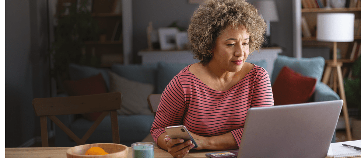 Smiling mid adult woman working on a computer while using mobile phone at home.