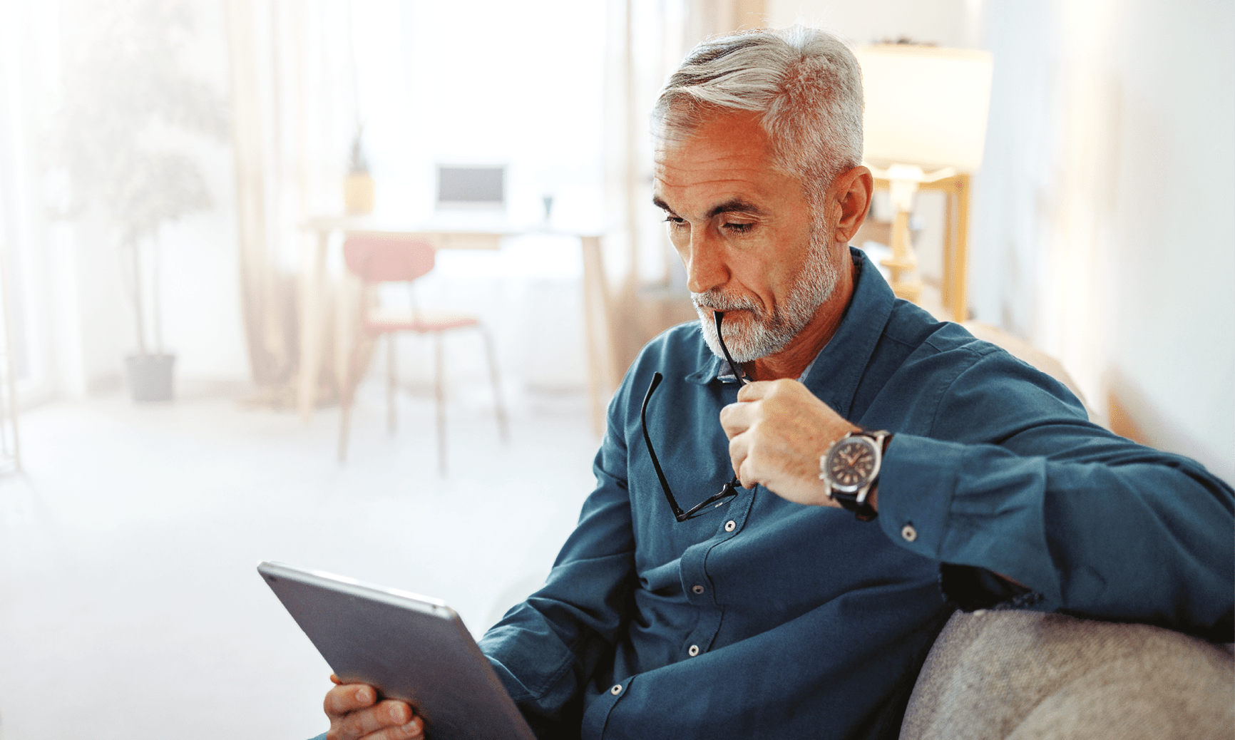 Casually clothed mature man using digital tablet while relaxing on a sofa at home