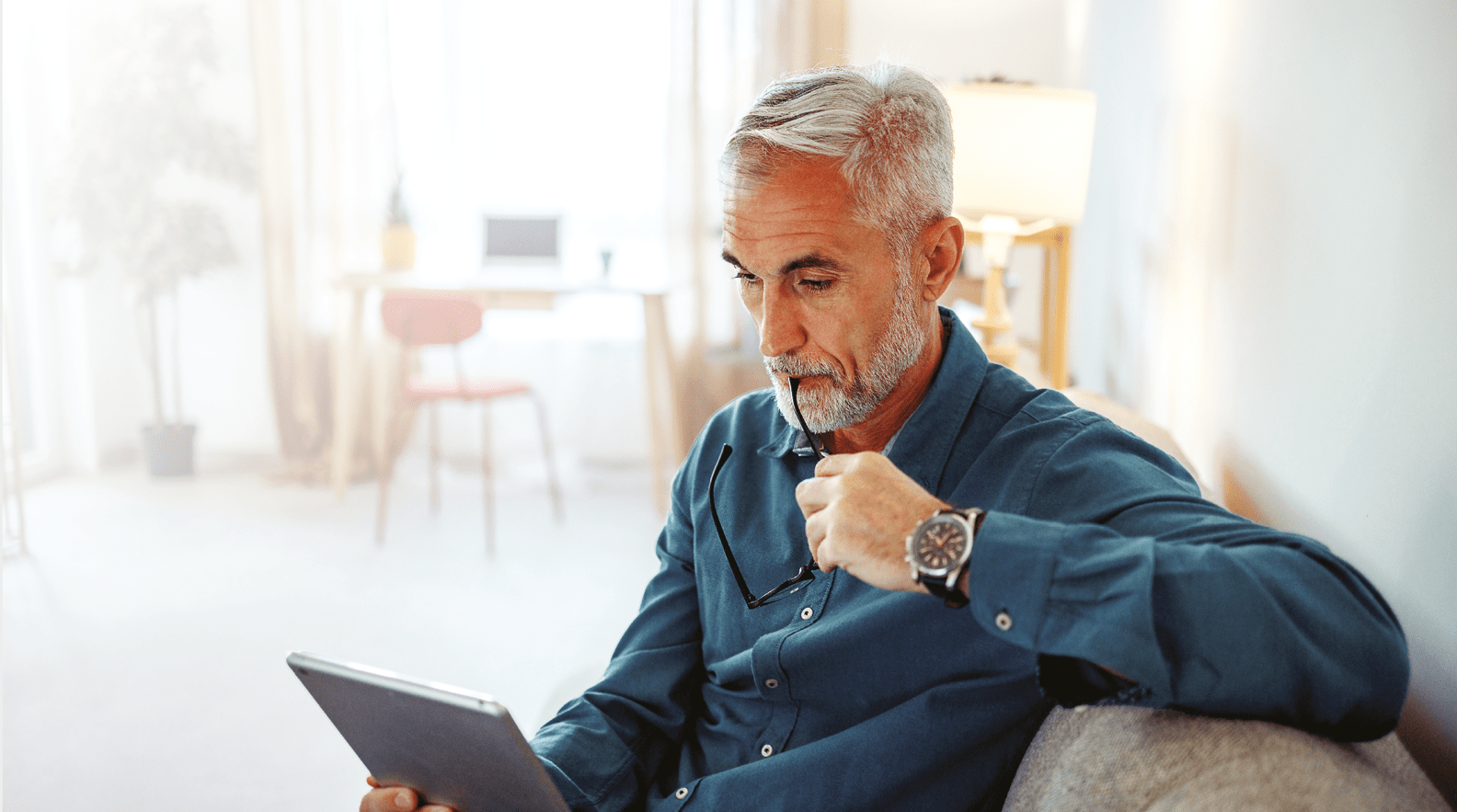 Casually clothed mature man using digital tablet while relaxing on a sofa at home