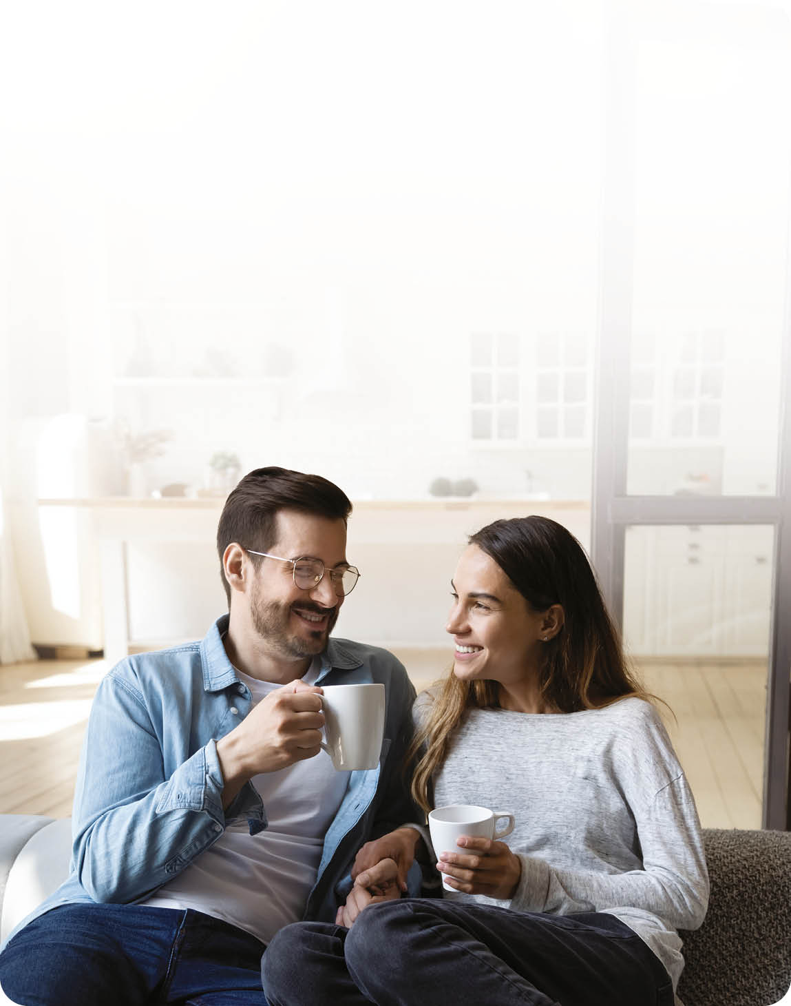 Happy young couple husband and wife relaxing on sofa in modern home living room talking laughing holding cups drinking tea bonding enjoying pleasant conversation romantic leisure lifestyle at home