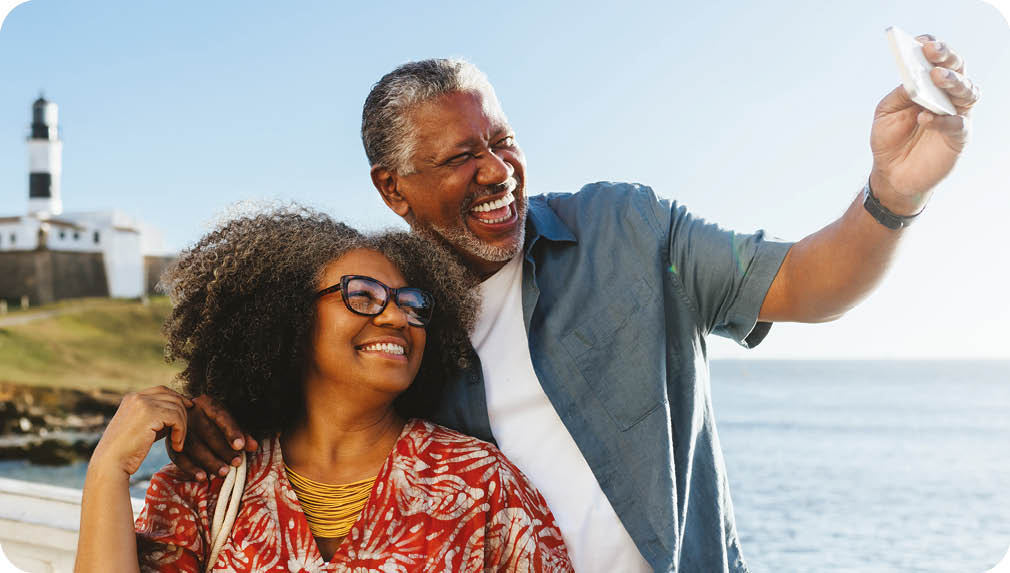 Happy elderly couple taking a selfie near the coast, with the sea and a lighthouse in the background. Retired Life at it's best.