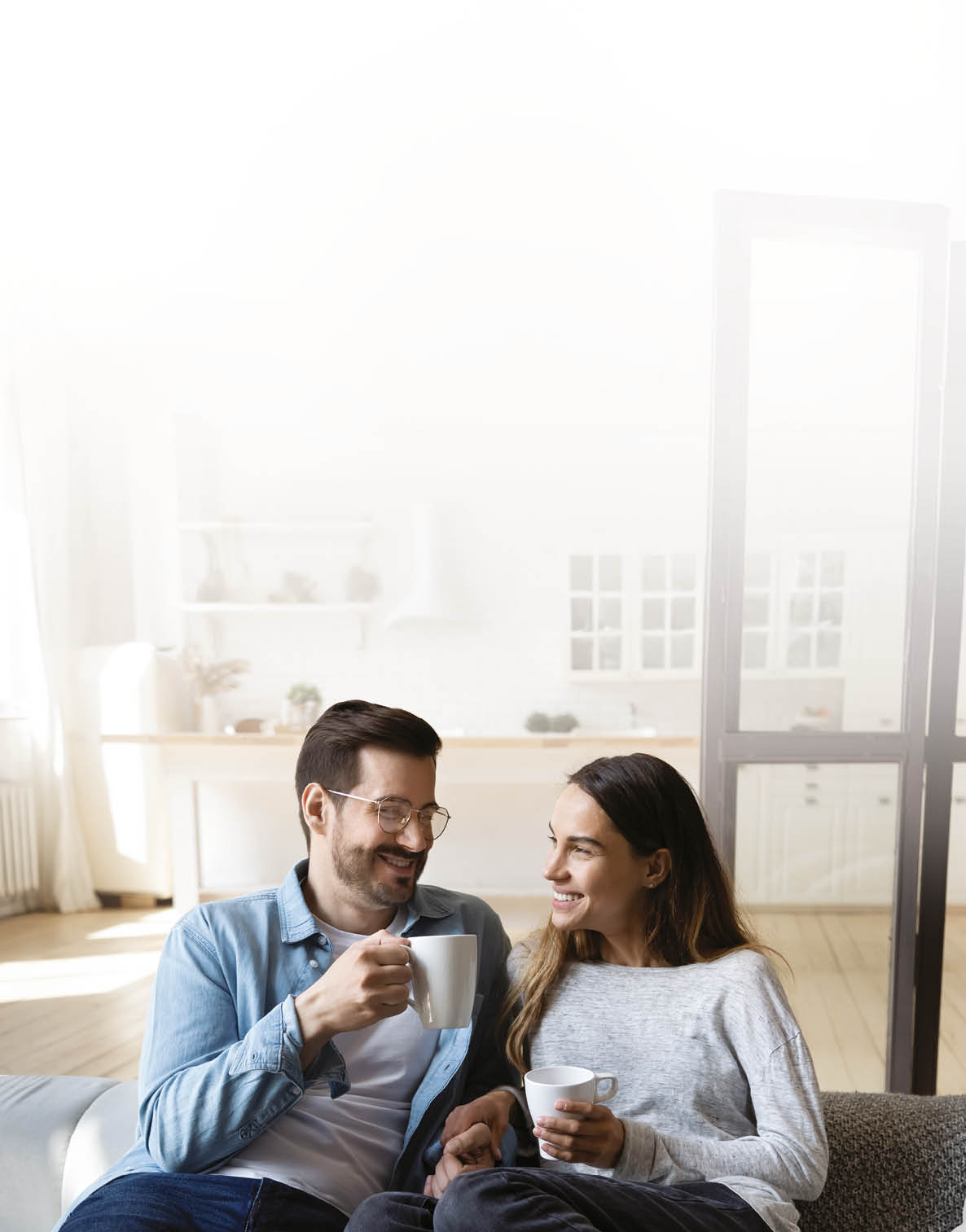 Happy young couple husband and wife relaxing on sofa in modern home living room talking laughing holding cups drinking tea bonding enjoying pleasant conversation romantic leisure lifestyle at home