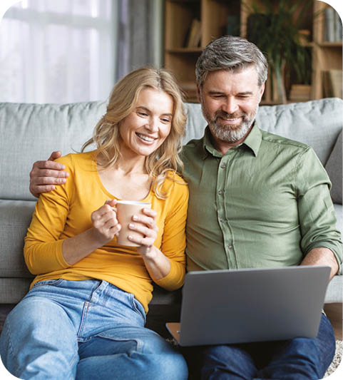 A man and woman sitting on a couch, the woman holding a cup and the man holding a laptop. Description generated by AI