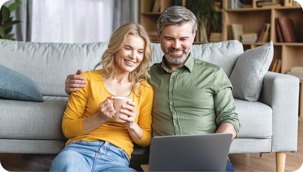 A man and woman sitting on a couch, the woman holding a cup and the man holding a laptop. Description generated by AI