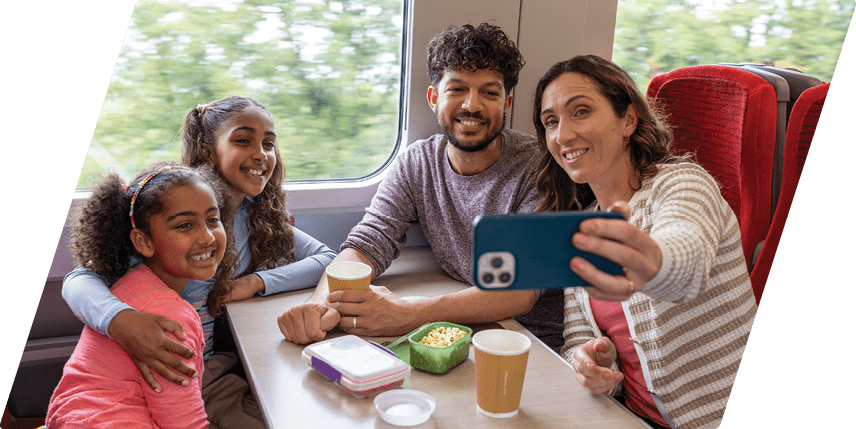 A wide shot of a family of four sitting around a table taking a selfie on a train during a journey from Newcastle to Edinburgh. They are all leaning in towards each other, smiling for the picture, the mother holding the mobile phone out. They are dressed in casual clothing and it is summertime. On the table there are various plastic containers of food and paper coffee cups. Videos similar to this scenario available.