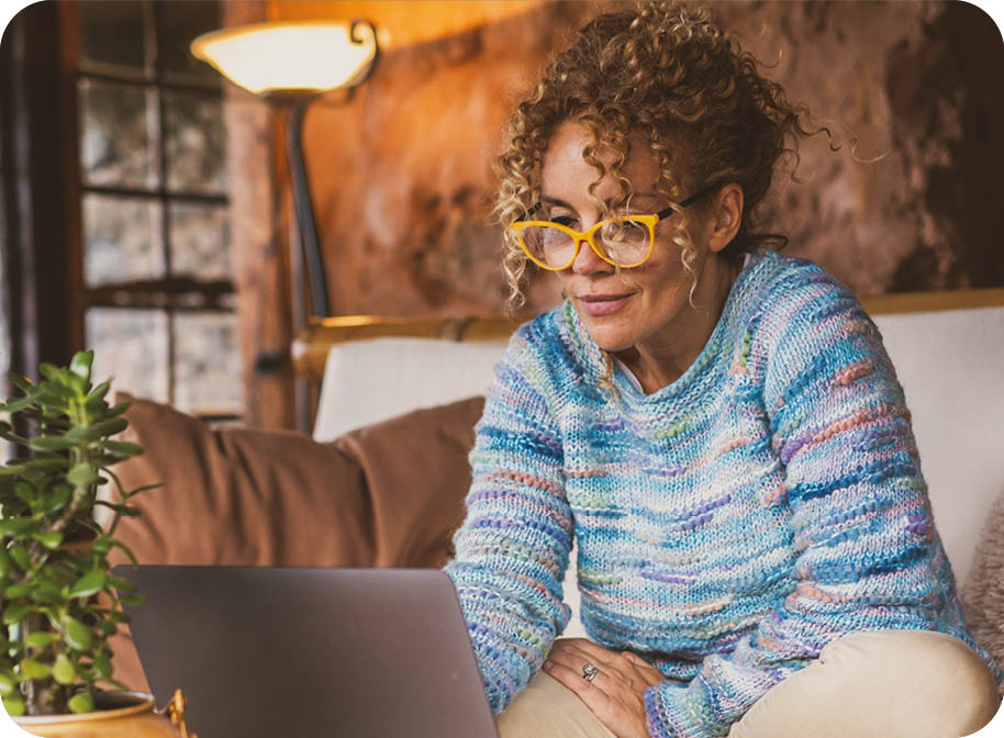 A woman wearing glasses and a sweater is sitting on a couch and using a laptop. AI generated content
