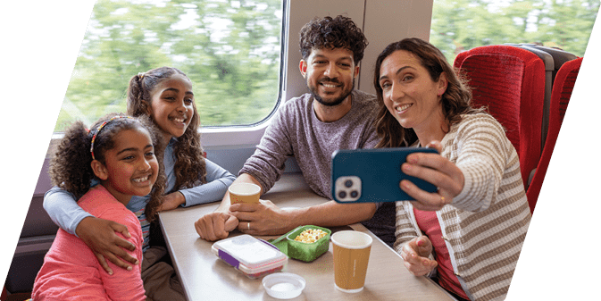 A wide shot of a family of four sitting around a table taking a selfie on a train during a journey from Newcastle to Edinburgh. They are all leaning in towards each other, smiling for the picture, the mother holding the mobile phone out. They are dressed in casual clothing and it is summertime. On the table there are various plastic containers of food and paper coffee cups. Videos similar to this scenario available.
