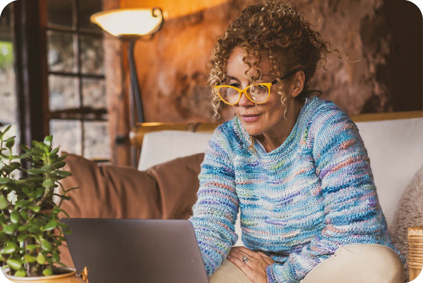 A woman wearing glasses and a sweater is sitting on a couch and using a laptop. AI generated content