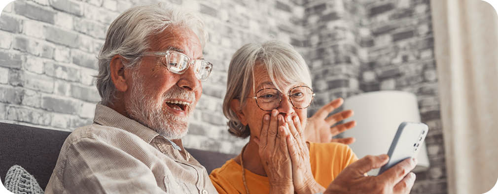 An elderly man and woman are sitting on a couch, looking at a cell phone. The woman is wearing glasses and has her mouth open, possibly reacting to something on the screen. AI generated content