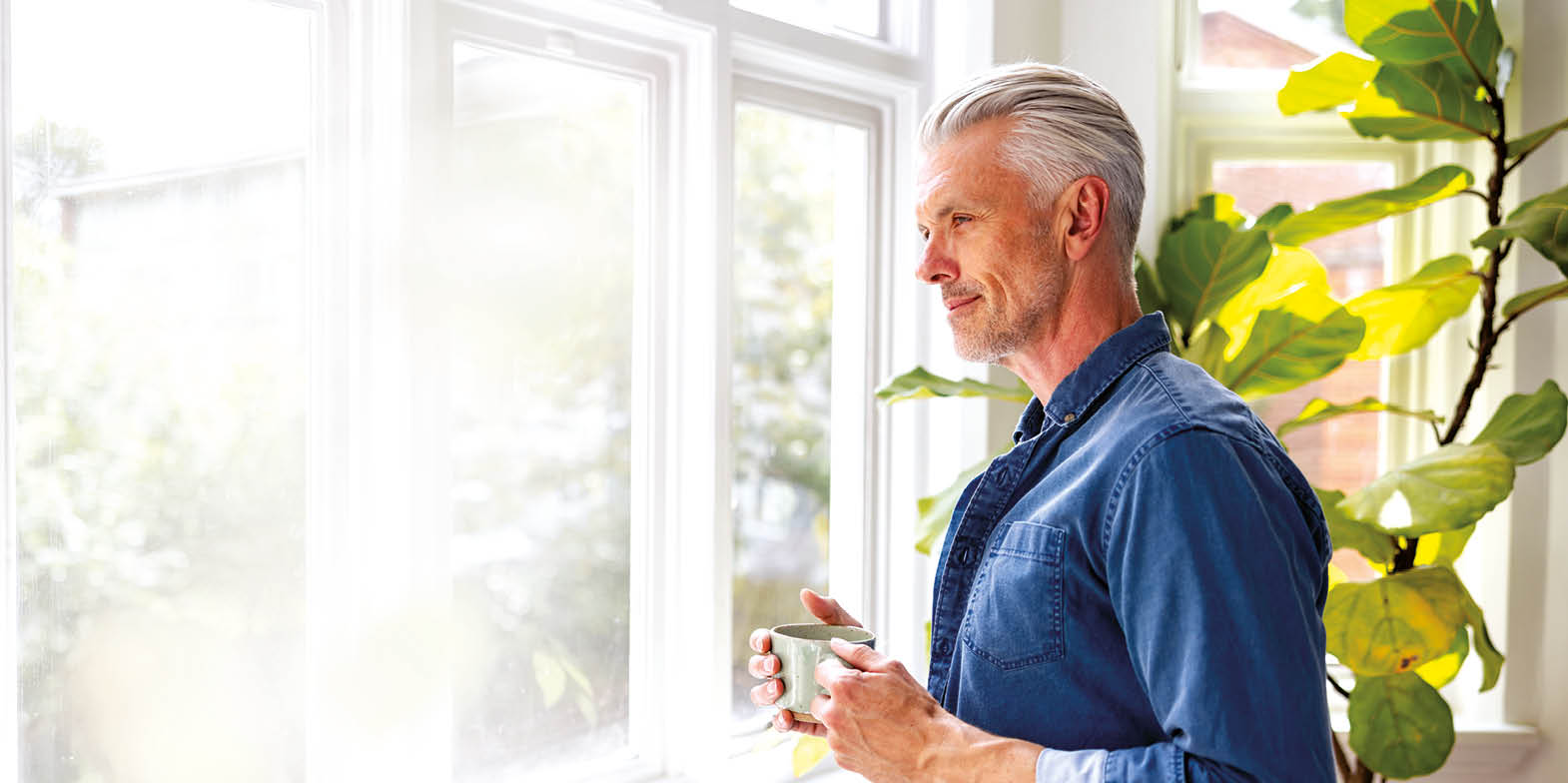 Happy man at home drinking a cup of coffee while looking through the window - domestic life concepts