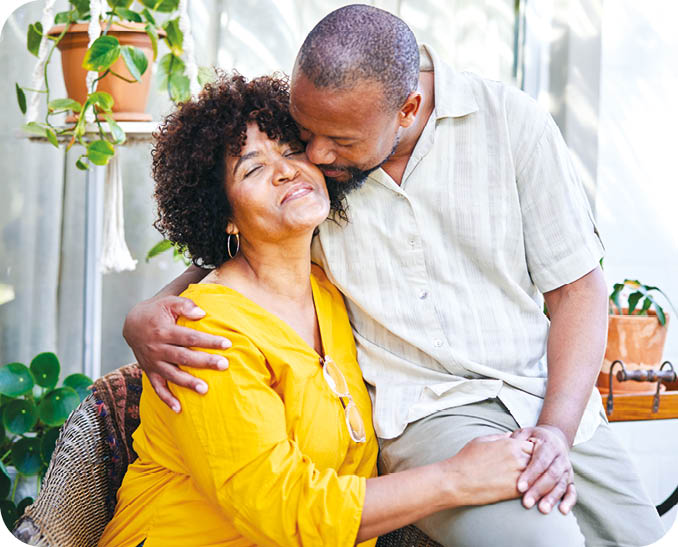 Loving mature man kissing his smiling wife on the cheek while sitting together outside on their patio at home in summer