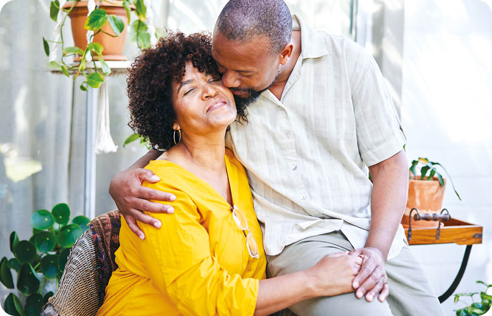 Loving mature man kissing his smiling wife on the cheek while sitting together outside on their patio at home in summer