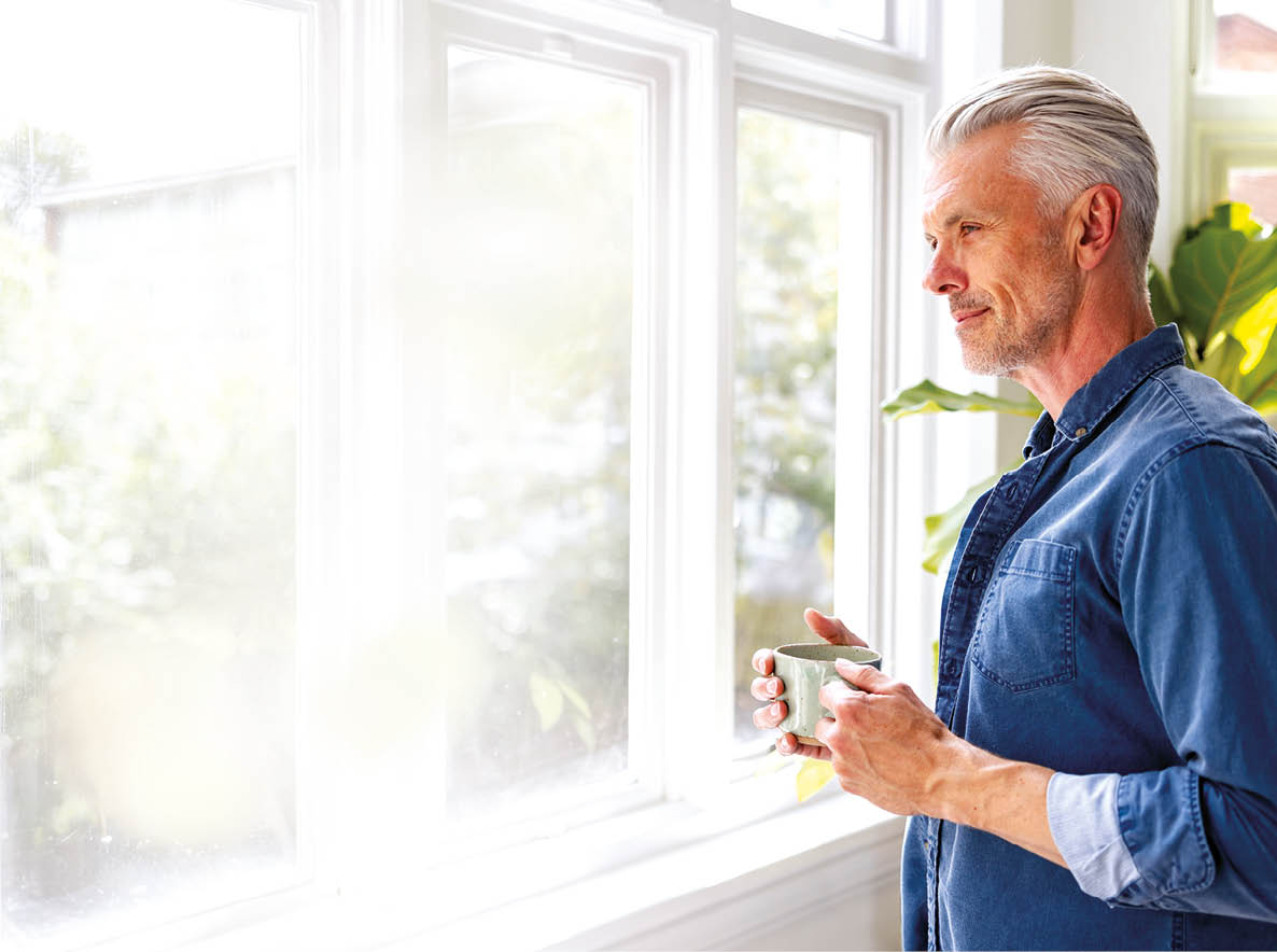 Happy man at home drinking a cup of coffee while looking through the window - domestic life concepts