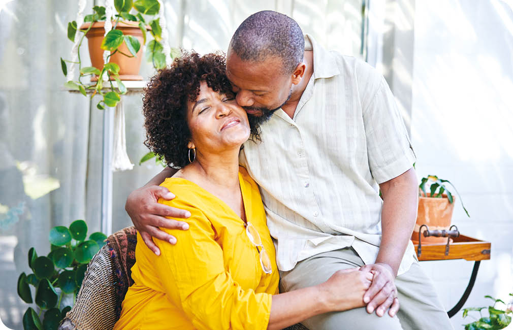 Loving mature man kissing his smiling wife on the cheek while sitting together outside on their patio at home in summer
