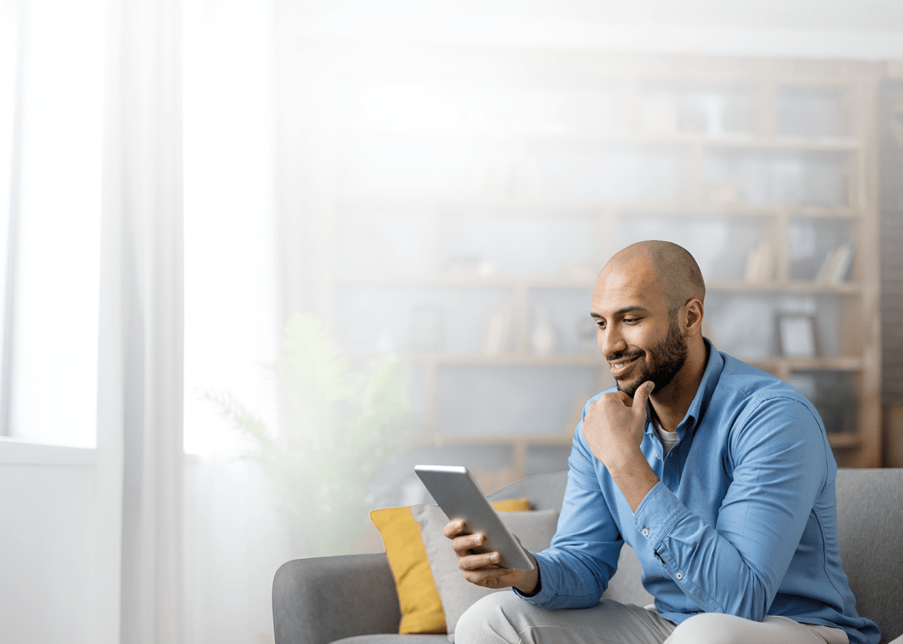 A person wearing a blue shirt is sitting on a gray couch in a bright room, smiling while holding and looking at a tablet. Description generated by AI