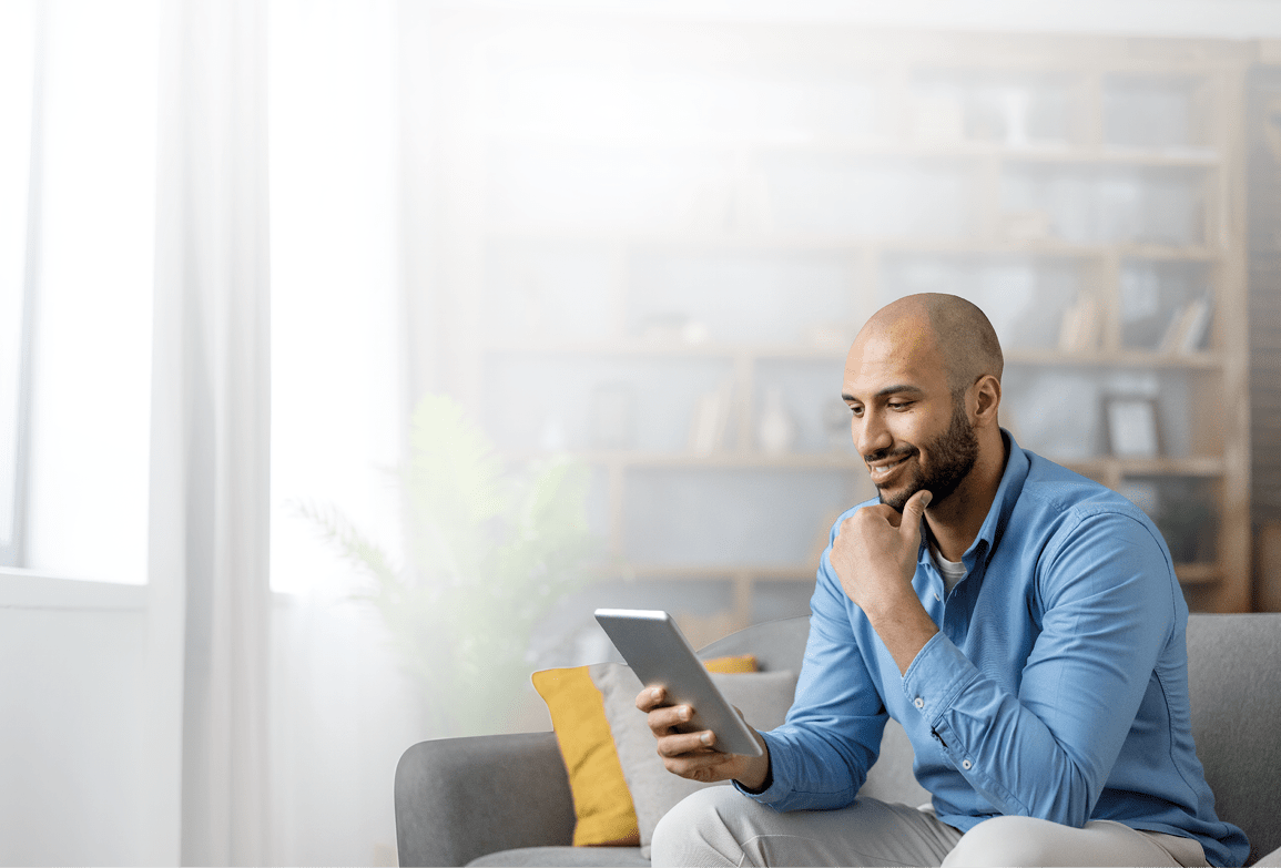A person wearing a blue shirt is sitting on a gray couch in a bright room, smiling while holding and looking at a tablet. Description generated by AI