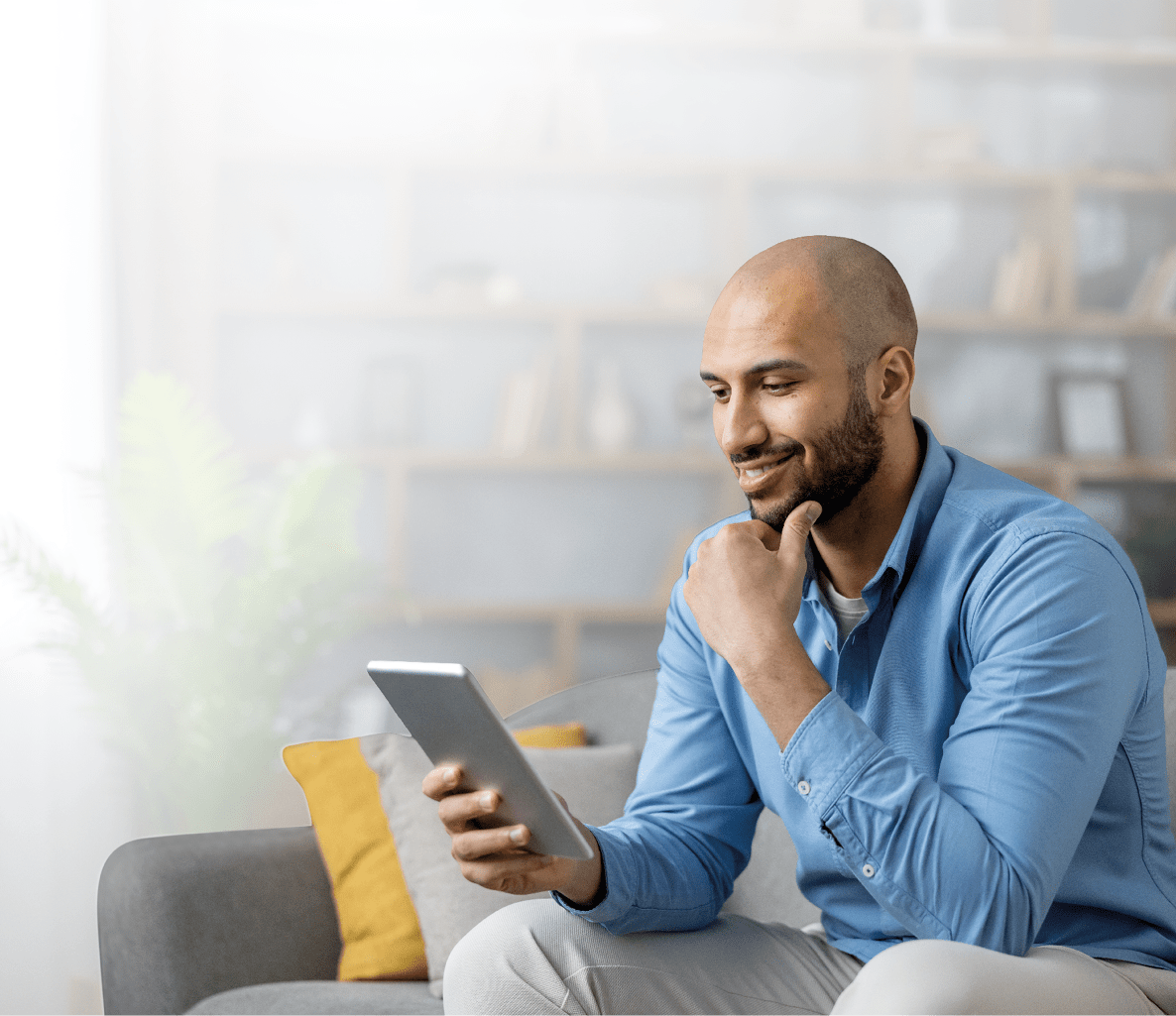A person wearing a blue shirt is sitting on a gray couch in a bright room, smiling while holding and looking at a tablet. Description generated by AI